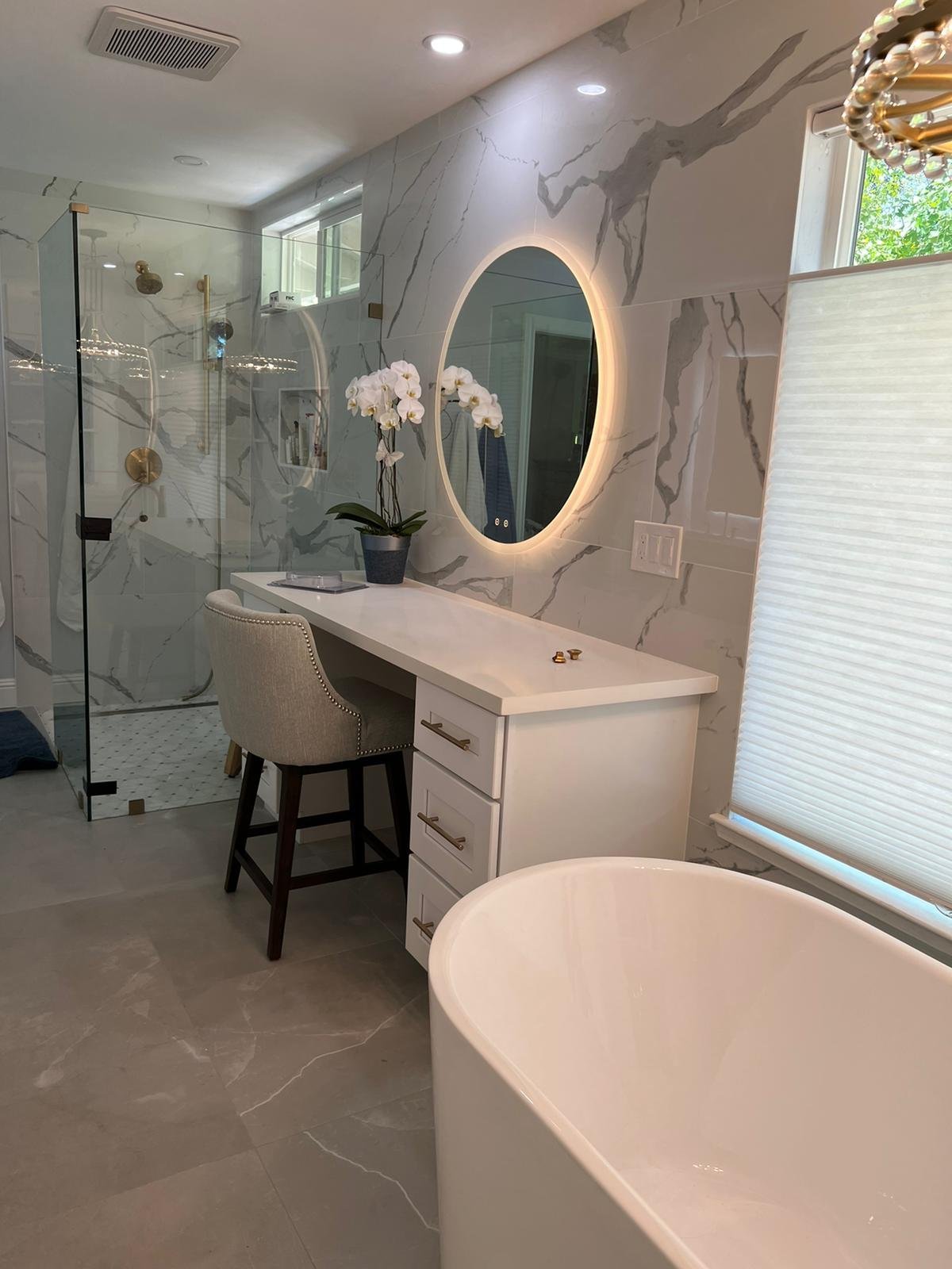 Bathroom makeup vanity with a round lit mirror beside a marble-look walk-in shower and freestanding tub