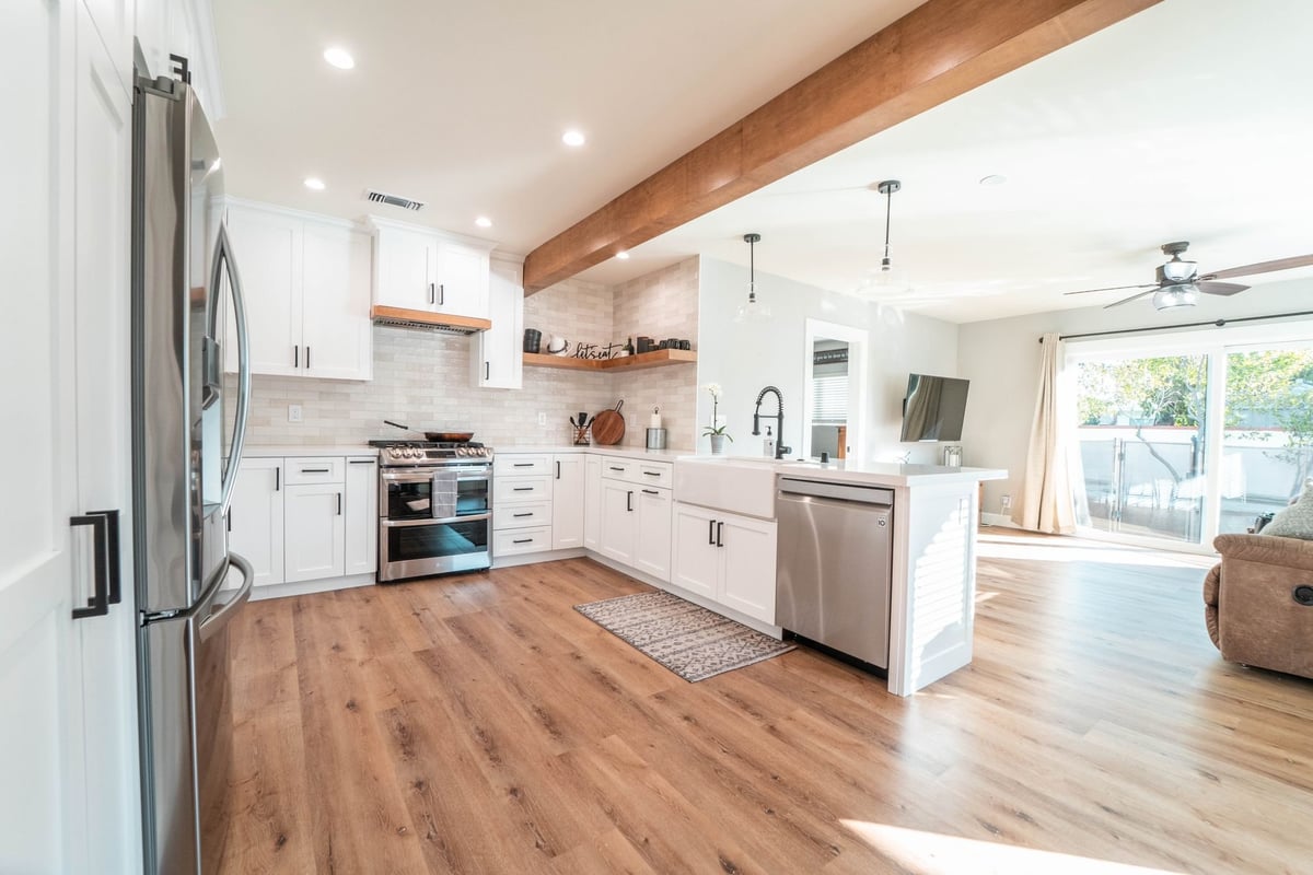 Open-concept remodeled kitchen with white shaker cabinets, wood-look flooring and an exposed wood beam opening to the living room
